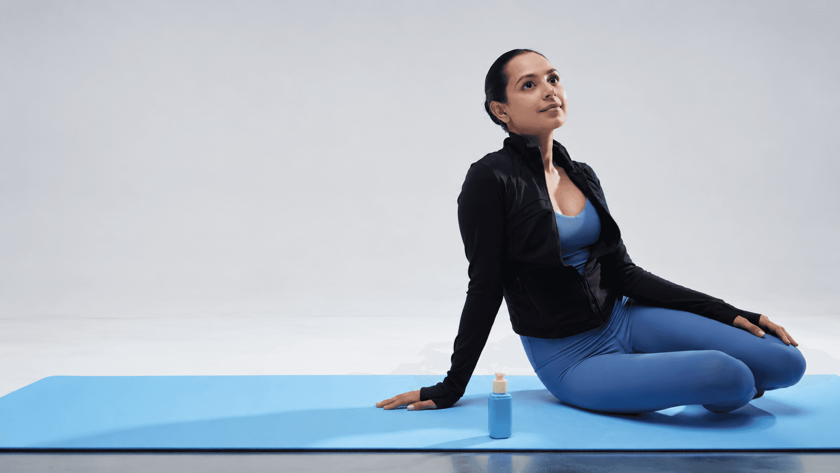 A woman pausing mid-stretch on a blue exercise mat, a bottle of Felihz mist beside her